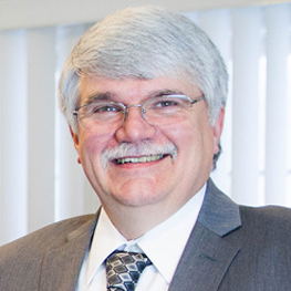 Headshot of Danny Desrosiers, with gray hair and mustache, glasses, smiling and wearing a suit jacket and tie